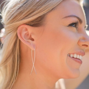 May include: A woman wearing silver, thin, long, crossed earrings. The earrings are simple and delicate.