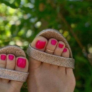 May include: A pair of glittery gold slide sandals with a thick strap across the toes. The toes are painted with a bright hot pink nail polish, and a thin, simple gold-toned metal toe ring is worn on the second toe of the right foot.