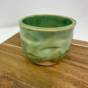 May include: A handmade, green ceramic bowl with a textured exterior. The bowl has a glossy finish and a rounded shape. The interior is a lighter shade of green. The bowl is sitting on a wooden surface.