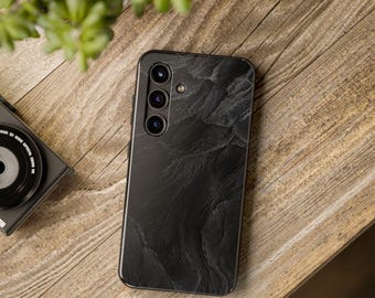 May include: A smartphone case featuring a dark gray and black textured stone pattern. The case has a vertical camera array with three circular lenses and a small flash unit positioned on the upper left side of the back panel.