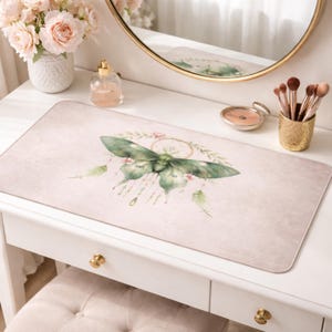 May include: A rectangular desk mat with a watercolor design of a green moth with a dreamcatcher and floral accents. The mat has a soft pink background and is placed on a white vanity table.