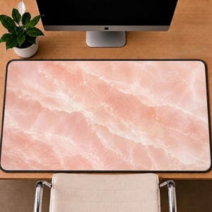 May include: A rectangular desk mat featuring a soft pink marble pattern with white veining throughout. The mat has a thin black border and a smooth surface, designed to sit flat on a workspace surface beneath a computer monitor and keyboard.