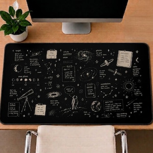 May include: A rectangular black desk mat featuring white illustrations of space, including an astronaut, planets, galaxies, a telescope, and moon phases. The design includes handwritten-style text notes about astronomy, physics equations, and scientific observations.