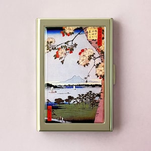 May include: A silver metal business card holder with a Japanese landscape painting. The artwork depicts a lake scene with a mountain in the background, cherry blossoms, and small boats. The holder has a rectangular shape.