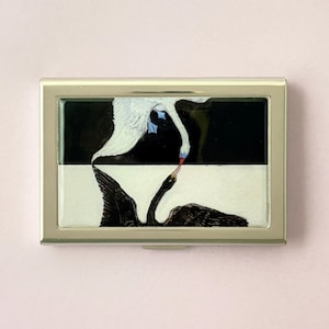 May include: A silver metal business card holder with a black and white swan design. The artwork features a white swan above a black swan, with the swans' beaks touching. The background is divided into black and white sections.