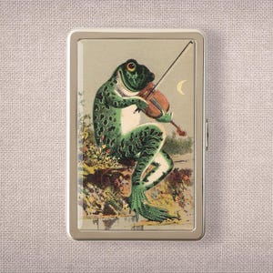 May include: A rectangular metal case featuring a printed illustration of a green spotted frog playing a brown violin. The frog is positioned on a textured ground with foliage, set against a light background with a crescent moon in the upper right corner.