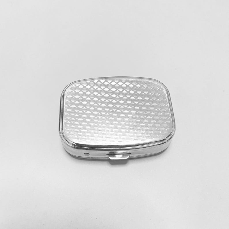 May include: A silver pill box with a patterned lid. The lid has a diamond pattern.