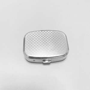May include: A silver pill box with a patterned lid. The lid has a diamond pattern.