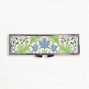 Op de afbeelding: Een zilveren pillendoosje met een scharnierend deksel. Het doosje is versierd met een bloemmotief in tinten groen, blauw en rood.