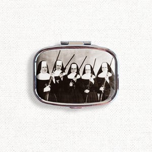 May include: A vintage-style compact mirror featuring a black and white photograph of five nuns holding rifles. The nuns are wearing traditional habits and are looking directly at the camera.