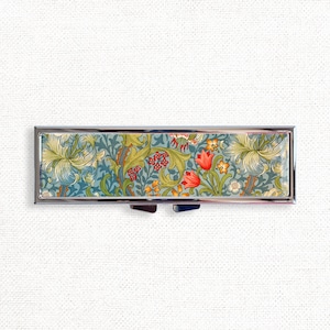 May include: A silver pill box with a floral design. The design features red, yellow, and green flowers on a blue background.