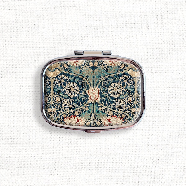 May include: A rectangular, silver-framed pill box with a floral pattern in teal, cream, and red. The design features stylized flowers and foliage against a dark teal background. The pill box is closed and sits on a white surface.
