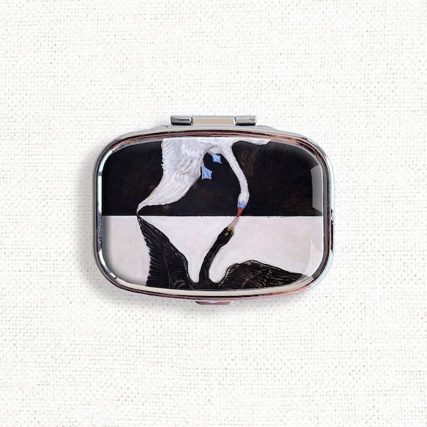 May include: A silver pill box with a black and white swan design. The artwork depicts a white swan above a black swan, with their beaks touching. The background is divided into black and white sections.