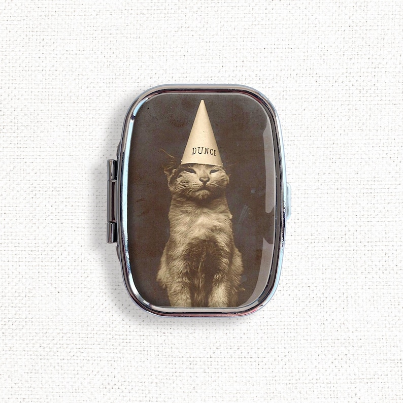 May include: A silver compact mirror with a vintage photo of a cat wearing a dunce cap. The cat is looking at the camera with a serious expression. The text on the cap reads "DUNCE".
