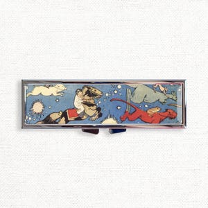 May include: A silver pill box with a colorful illustration of a child riding a horse, a kangaroo, a frog, and a red dog. The illustration is set against a blue background with white stars.