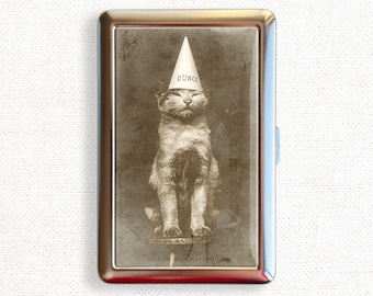 May include: A vintage black and white photograph of a cat wearing a dunce cap. The cat is sitting on a stool and looking directly at the camera. The image is printed on a silver metal cigarette case.