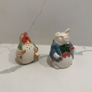 May include: A set of ceramic salt and pepper shakers featuring a rooster and a pig. The rooster is red, green, and white with a brown beak. The pig is white with a blue jacket, green bow tie, and red apple.