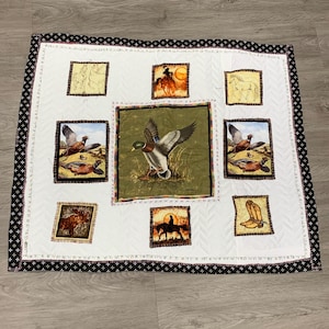 Puede incluir: Una colcha blanca con un borde negro y nueve cuadrados. Cada cuadrado presenta una imagen diferente de temática de caza o del oeste, incluyendo un pato, un faisán, un vaquero y un par de botas.