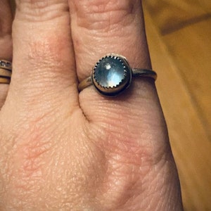 Op de afbeelding: Een zilveren ring met een ronde, iriserende maansteen in een bezel-setting. De ring is op een vinger.