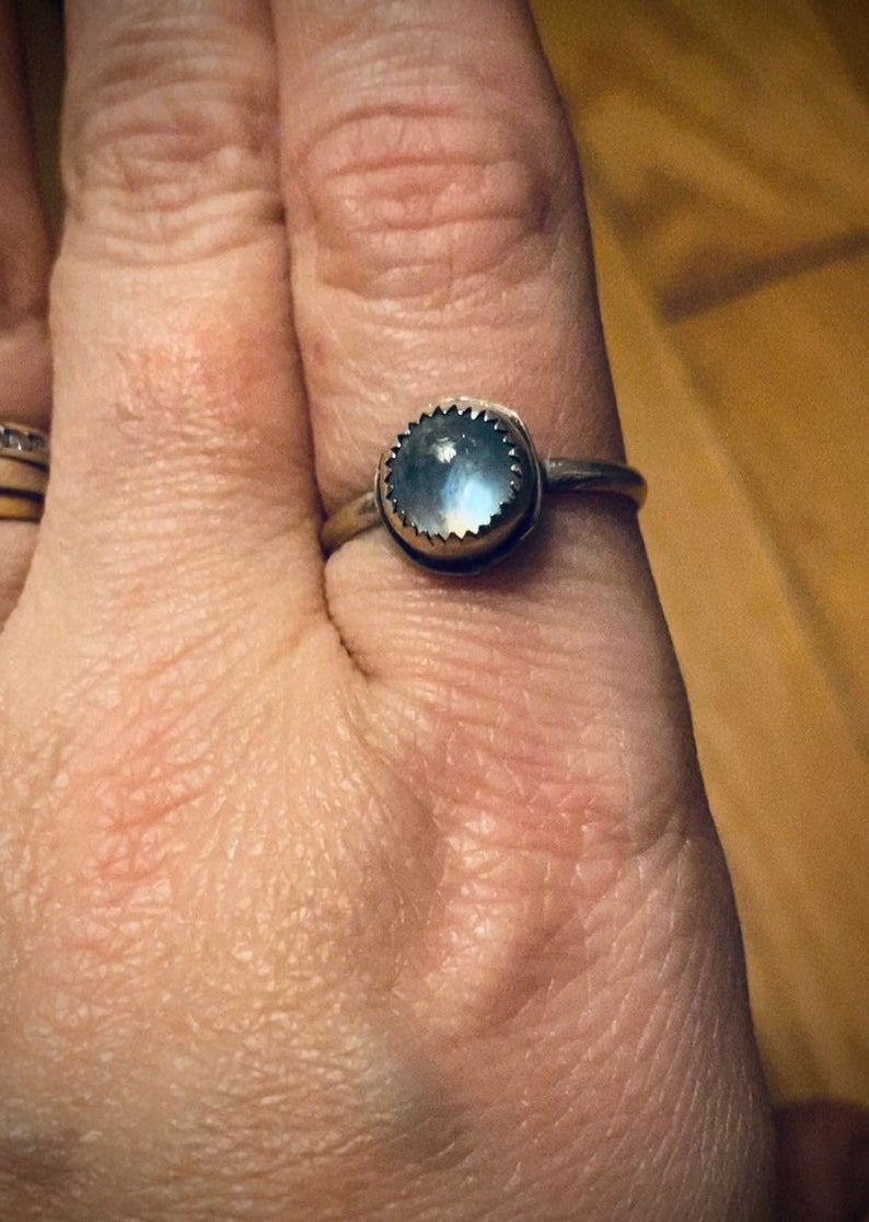 Op de afbeelding: Een zilveren ring met een ronde, iriserende maansteen in een bezel-setting. De ring is op een vinger.