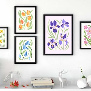 Puede incluir: Una colección de láminas artísticas florales enmarcadas. Las láminas presentan ilustraciones estilizadas de varias flores, como tulipanes, iris y narcisos, en colores vibrantes. Los marcos son negros y la obra de arte se exhibe sobre una pared blanca.