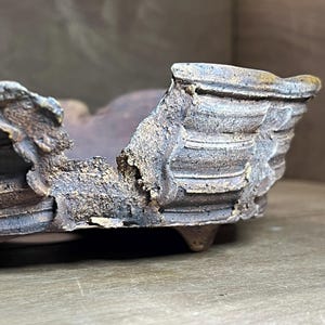 May include: A low-profile ceramic bonsai pot with a dark, weathered, and textured finish. The exterior features horizontal ridges and a rugged, uneven rim. The vessel is supported by small, short feet, showcasing a raw, earthy aesthetic with visible mineral deposits and a matte, multi-tonal brown and charcoal gray surface.