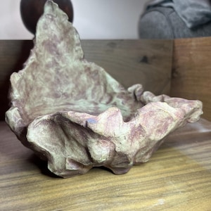 May include: A handcrafted, organic-shaped bowl with a textured surface. The bowl features a blend of muted pink and beige tones, creating a natural, earthy aesthetic. The bowl is displayed on a wooden surface.