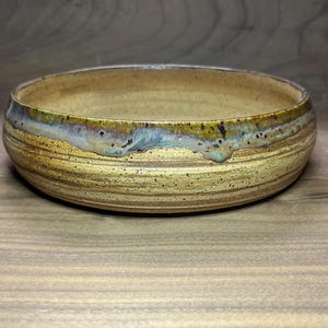 May include: A handmade ceramic bowl with a wide, shallow design. The bowl features a warm, earthy color palette with a speckled, textured surface. The rim has a unique glaze with shades of blue, green, and gold. The bowl is approximately 2 inches tall and 8 inches in diameter.