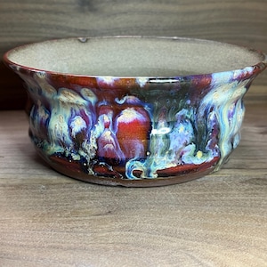 May include: A handmade ceramic bowl with a unique glaze. The bowl features a rim with a light tan color, and the exterior is a blend of red, blue, and green hues. The bowl's design is organic and textured, with a glossy finish.