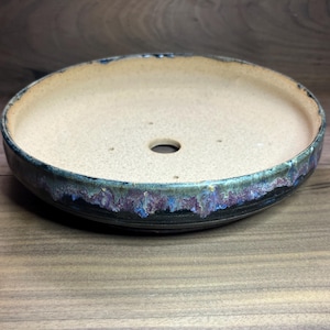 May include: A round, ceramic bonsai pot with a speckled beige interior and a dark, textured exterior rim. The rim features a blend of blue, purple, and black glaze. A single drainage hole is visible in the center of the pot.