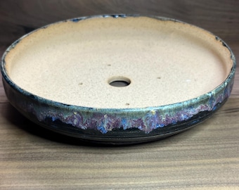May include: A round, ceramic bonsai pot with a speckled beige interior and a dark, textured exterior rim. The rim features a blend of blue, purple, and black glaze. A single drainage hole is visible in the center of the pot.