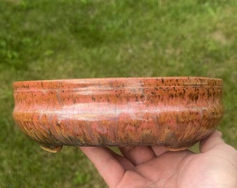May include: A handmade, low-profile ceramic bowl with a reddish-orange glaze. The bowl has a wide, shallow shape and sits on three small feet. The glaze has a mottled appearance with hints of brown and gold. The bowl is held in a hand.