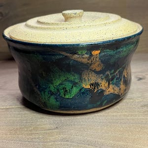 May include: A handmade ceramic lidded bowl with a speckled beige lid and a dark blue, green, and orange glazed body. The bowl has a rounded shape and sits on a wooden surface. The lid has a small knob for easy handling.