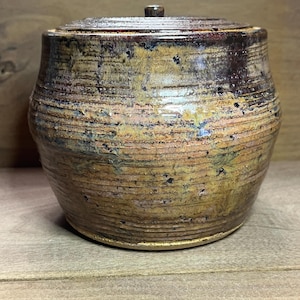May include: A handmade ceramic jar with a lid. The jar has a round body and a lid with a small knob. The exterior features horizontal ridges and a blend of brown, yellow, and black colors. The jar is sitting on a wooden surface.