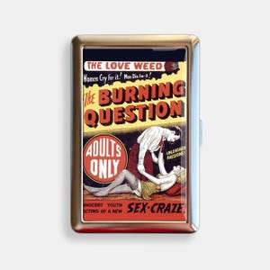 May include: A silver cigarette case with a retro graphic. The graphic includes the text "THE LOVE WEED" and "The BURNING QUESTION", alongside an illustration of two people. The case also features the text "ADULTS ONLY" and "SEX-CRAZE".
