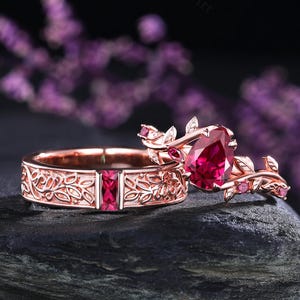 Può includere: Due anelli in oro rosa. Uno presenta una grande gemma rossa a forma di cuore, l'altro ha una gemma rossa rettangolare. Entrambi gli anelli hanno un design floreale. Gli anelli sono esposti su una superficie scura.