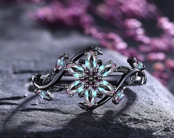 Puede incluir: Un anillo de metal negro con un grupo central en forma de flor de piedras iridiscentes de talla marquesa. La banda está diseñada con metal en forma de enredadera que se extiende hacia afuera, incorporando pequeñas piedras iridiscentes a juego en los lados.