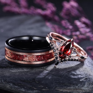 Può includere: Un set di fedi nuziali abbinate con una fascia nera con intarsio rosso e oro e un anello di fidanzamento in oro rosa con una pietra rossa taglio marquise e pietre nere. Gli anelli sono esposti su una pietra grigio scuro.