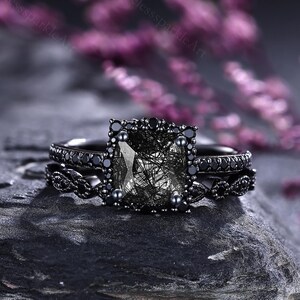 May include: A black gemstone ring set with a square-cut central stone and smaller surrounding stones. The ring rests on a dark grey rock, with blurred purple flowers in the background. The ring has a matching band with detailed design.