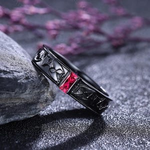 以下が含まれることがあります： 正方形の赤い宝石が付いた黒い指輪。指輪は、花柄と幾何学模様の装飾的なデザインです。指輪は、背景に紫色の花がある、濃い灰色の石の表面に表示されています。