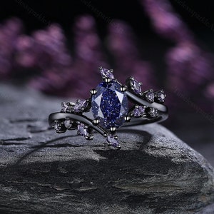 Könnte beinhalten: Ein schwarzer Ring mit einem großen, ovalen, glitzernden lila Edelstein, der von kleineren lila Edelsteinen umgeben ist. Der Ring hat ein zartes, gedrehtes Band mit einem Blattdesign.