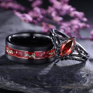 Puede incluir: Un juego de anillos de boda negros. Uno tiene una incrustación roja y dorada, el otro una piedra roja en forma de marquesa y detalles negros. Los anillos se exhiben sobre una piedra gris oscura. Par a juego.
