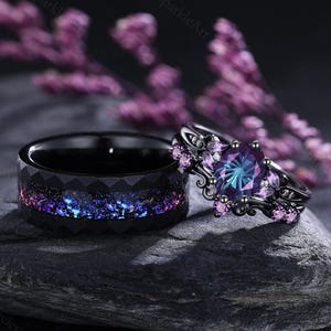 Può includere: Due anelli, uno nero con un design sfaccettato e un intarsio galattico viola e blu. L'altro anello è nero con una grande gemma ovale viola-blu e gemme rosa più piccole, su una fascia decorativa. Su una pietra grigio scuro.