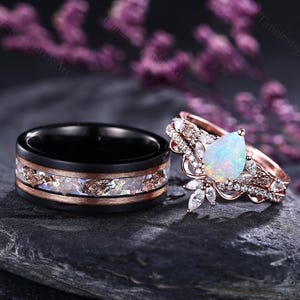 May include: A black wedding band with a copper-colored inlay and a rose gold engagement ring with a pear-shaped opal center stone and diamond accents. The rings are displayed on a dark gray stone.
