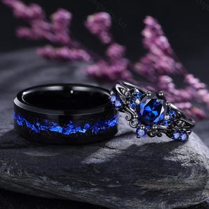Può includere: Un set di fedi nuziali nere. La fede ha un intarsio blu, mentre l'anello di fidanzamento presenta una grande pietra preziosa blu ovale e pietre blu più piccole. Gli anelli sono esposti su una pietra scura. Un set abbinato.