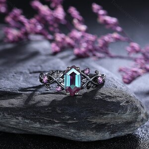 May include: A black ring with a hexagonal, colour-changing gemstone. The ring features a leaf design with small pink gemstones. The ring is set on a dark grey stone, with blurred purple flowers in the background.