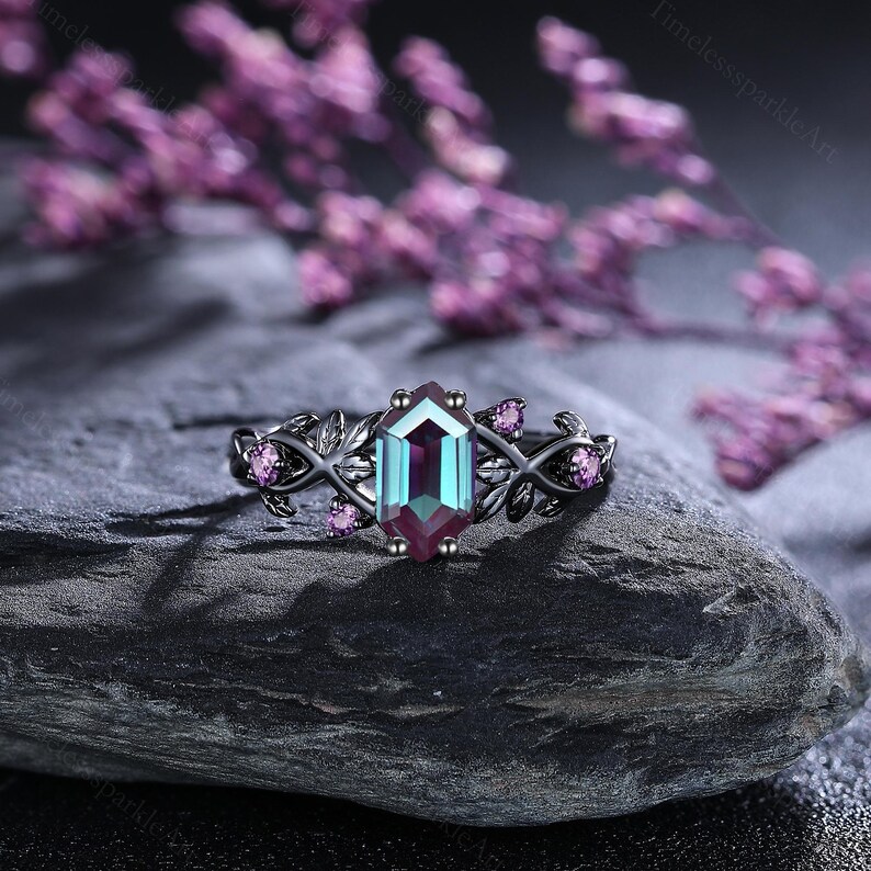 May include: A black ring with a hexagonal, colour-changing gemstone. The ring features a leaf design with small pink gemstones. The ring is set on a dark grey stone, with blurred purple flowers in the background.