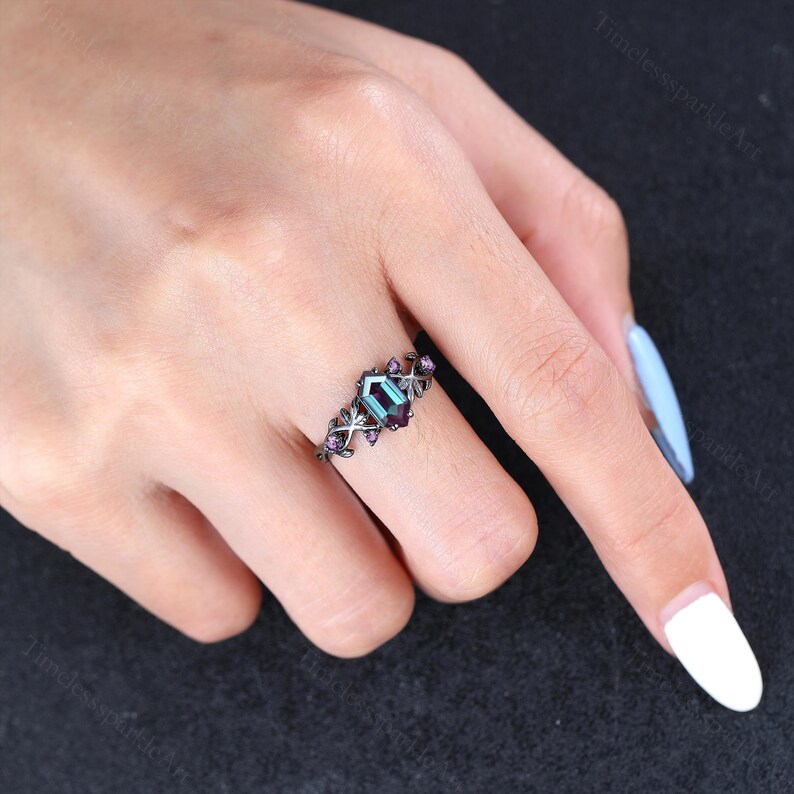 May include: A black metal ring featuring a hexagonal teal gemstone. The ring is embellished with small, purple gemstones and decorative filigree. The ring is on a person's finger.