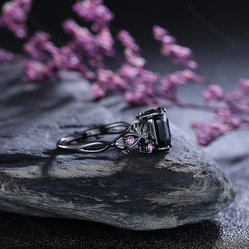 May include: A black ring featuring a large, dark square-cut stone and small pink accent stones. The ring has an intricate, twisted band design. The ring is set on a dark, textured surface with blurred purple flowers in the background.