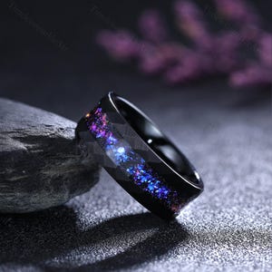 May include: Black ring with a galaxy design. The ring has a dark band with a textured pattern and a vibrant inlay of purple, blue, and pink colours, resembling a starry night. A unique piece of jewellery.